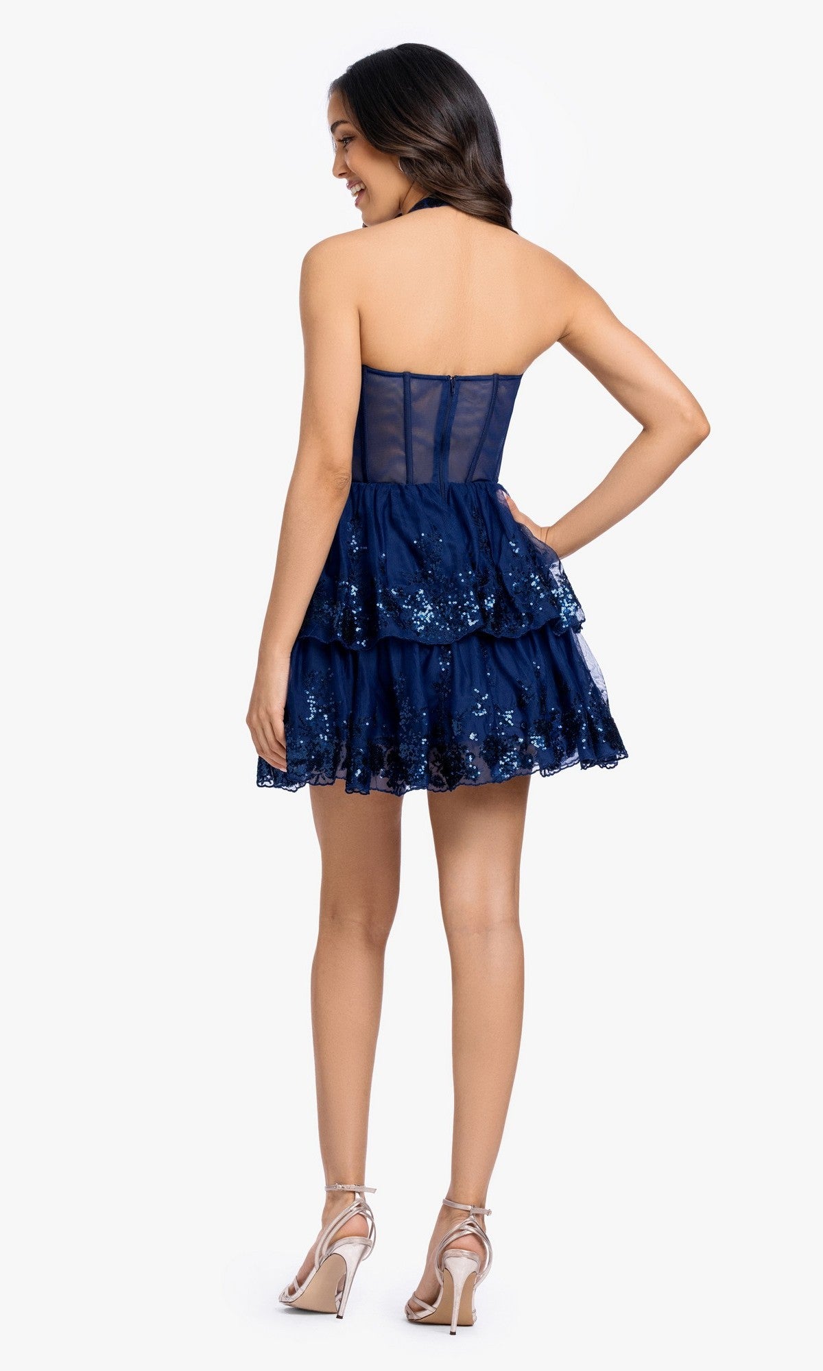 A woman stands facing away, wearing the Periwinkle Short Prom Dress: Blondie Nites 5702BN, a strapless sequin cocktail dress with tiered layers, paired with silver high-heeled sandals.